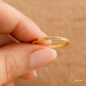 May include: A gold ring with the inscription "Leo & Bianca". The ring is held in a hand against a neutral background. The ring is a simple band design.