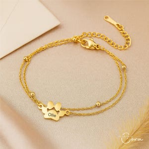 Puede incluir: Una pulsera dorada de doble cadena con un dije de huella de pata. El dije está grabado con el nombre "Ollie". La pulsera tiene un cierre de mosquetón y pequeños detalles de bolas.