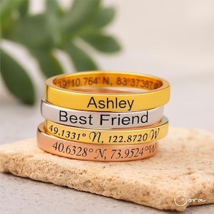 Puede incluir: Una pila de cuatro anillos en oro, plata y oro rosa. El anillo superior es dorado y dice "Ashley". El segundo anillo es plateado y dice "Best Friend". El tercer anillo es dorado y tiene coordenadas. El anillo inferior es de oro rosa y tiene coordenadas.