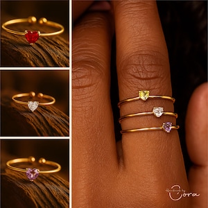 May include: Gold rings with heart-shaped gemstones in red, white, and purple. The rings are displayed on a finger, highlighting their delicate design. The gemstones are the focal point, adding a pop of colour.