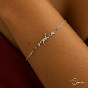 May include: A delicate silver bracelet with the name "Sophia" in cursive script. The bracelet is a fine chain with the name as a central design element. The bracelet is shown on a person's arm.