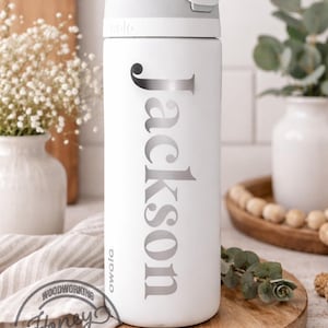 May include: White Owala water bottle with a gray lid and the name "Jackson" in silver lettering. The bottle is on a wooden tray, with a white background and decorative elements.