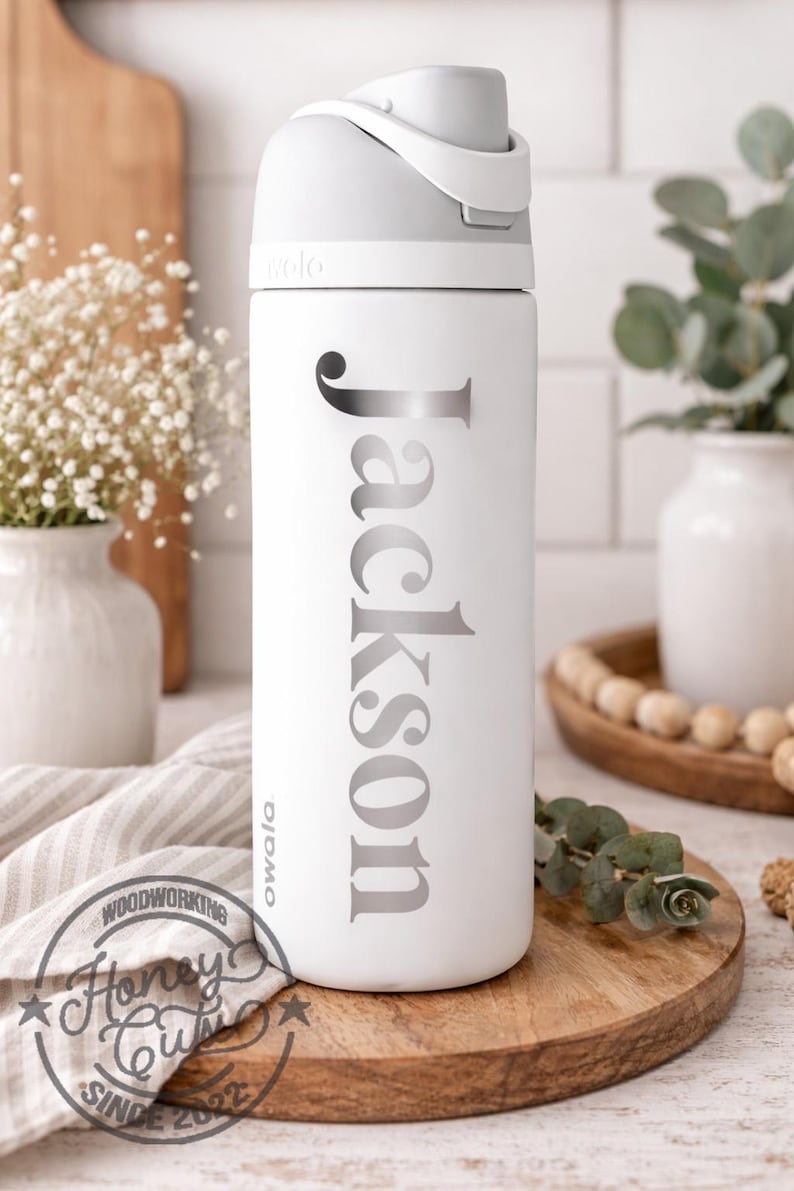 May include: White Owala water bottle with a gray lid and the name "Jackson" in silver lettering. The bottle is on a wooden tray, with a white background and decorative elements.