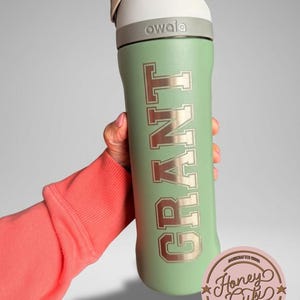 May include: A mint green Owala water bottle with the name "GRANT" in a collegiate font. The bottle has a white and gray lid. A pink and brown Honey Cubi sticker is in the lower right corner.