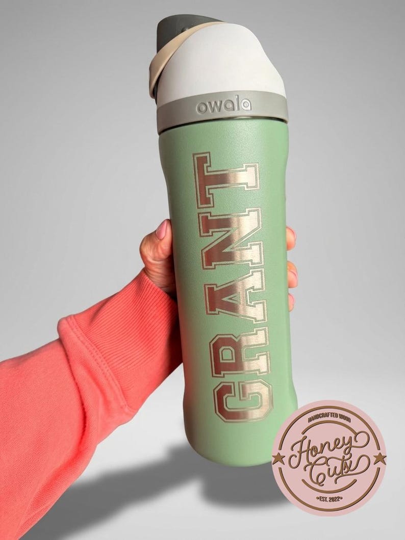 May include: A mint green Owala water bottle with the name "GRANT" in a collegiate font. The bottle has a white and gray lid. A pink and brown Honey Cubi sticker is in the lower right corner.
