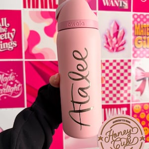 May include: A pink Owala water bottle with the name "Atalee" in gold script. The bottle has a flip-top lid and is held against a backdrop of pink and white patterned squares and inspirational quotes.