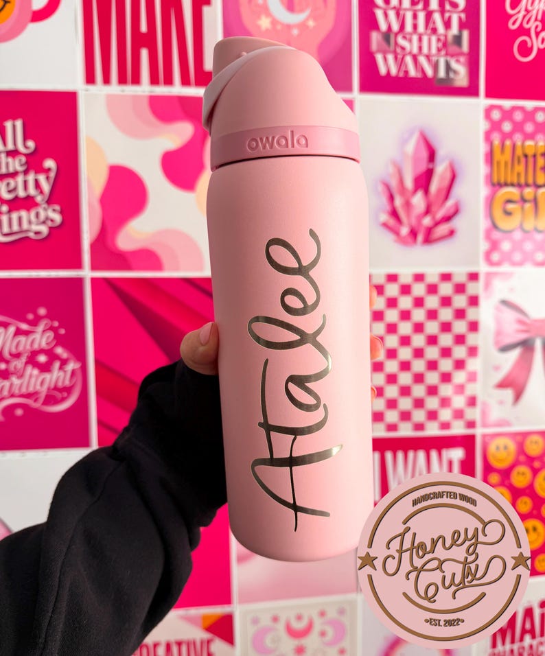 May include: A pink Owala water bottle with the name "Atalee" in gold script. The bottle has a flip-top lid and is held against a backdrop of pink and white patterned squares and inspirational quotes.