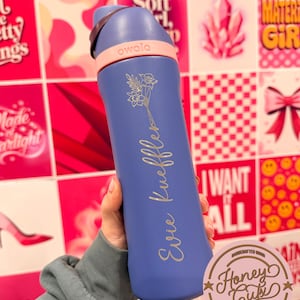 Puede incluir: Una botella de agua Owala azul con asa y detalles en rosa claro. La botella est&aacute; grabada con un dise&ntilde;o floral y el nombre "Evie Kueffler". El fondo presenta un collage de im&aacute;genes rosas y rojas.