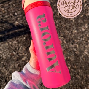 May include: A bright pink water bottle with the name "Aurora" in silver lettering. The bottle has a cream-colored top with a red band and the brand name "owala". A "Honey Cuts" logo is visible in the upper right corner.