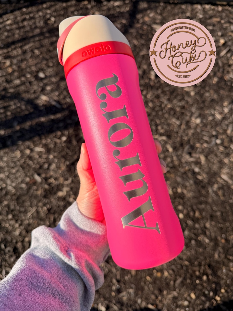 May include: A bright pink water bottle with the name "Aurora" in silver lettering. The bottle has a cream-colored top with a red band and the brand name "owala". A "Honey Cuts" logo is visible in the upper right corner.