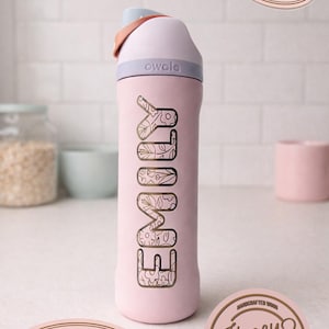 May include: A pink Owala water bottle with the name "EMILY" in a floral design. The bottle has a light pink lid and a handle. The image also includes a "Fast Shipping" sticker and a "Honey Cuts" logo.