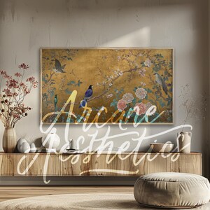 以下が含まれることがあります： 金色の背景と花柄の額縁付きの絵画。絵画には、ピンクの花が咲いた枝に止まっている青、黒、白の鳥がいくつか描かれている。