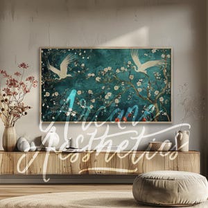 Op de afbeelding: Een tealblauwe canvas print met twee witte vogels die vliegen voor een bloeiende boom met witte bloemen. De print is ingelijst in een dun, licht houten frame en hangt aan een muur boven een houten plank.