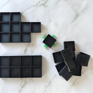 May include: Two black trays with 12 square compartments each, and a pile of black square boxes with the letter 'H' embossed on them.
