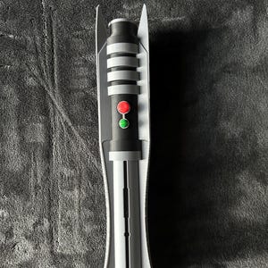 May include: A grey and black lightsaber prop with a red and green button. The hilt has silver bands and a white protective casing. The prop is designed to resemble a lightsaber from a science fiction franchise.