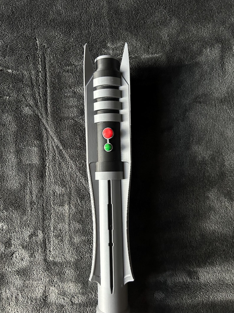 May include: A grey and black lightsaber prop with a red and green button. The hilt has silver bands and a white protective casing. The prop is designed to resemble a lightsaber from a science fiction franchise.