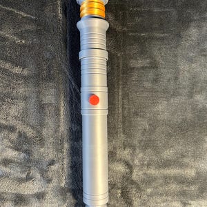 May include: A silver lightsaber prop with a gold and orange hilt and a red button. The lightsaber has a detailed design with ridges and a textured grip. The prop is designed to resemble a lightsaber from a science fiction franchise.