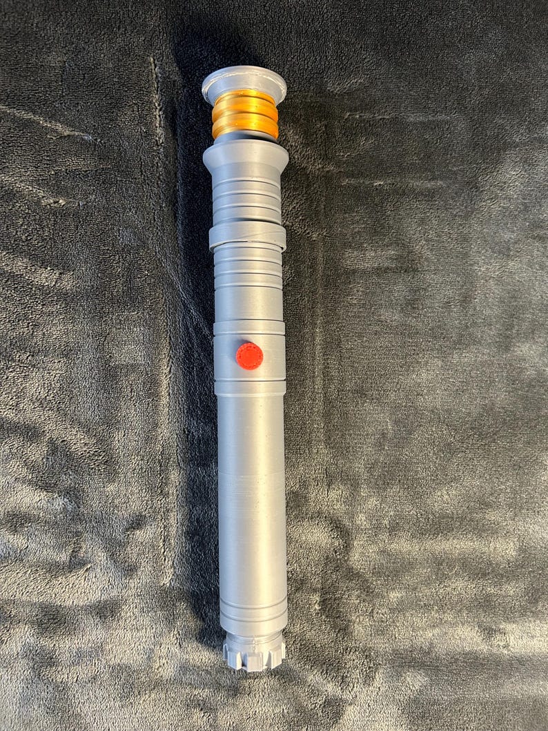 May include: A silver lightsaber prop with a gold and orange hilt and a red button. The lightsaber has a detailed design with ridges and a textured grip. The prop is designed to resemble a lightsaber from a science fiction franchise.