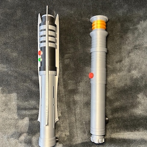 May include: Two toy lightsabers are shown on a dark grey surface. The lightsaber on the left is grey, black, and white with red and green buttons. The lightsaber on the right is grey with an orange top and a red button.