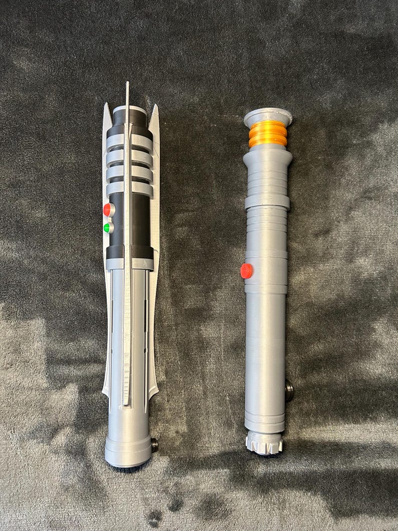 May include: Two toy lightsabers are shown on a dark grey surface. The lightsaber on the left is grey, black, and white with red and green buttons. The lightsaber on the right is grey with an orange top and a red button.