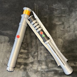 May include: Two toy lightsabers. One is silver with a red button and orange accents. The other is silver and black with a red and green button. The lightsabers are lying on a dark grey surface.