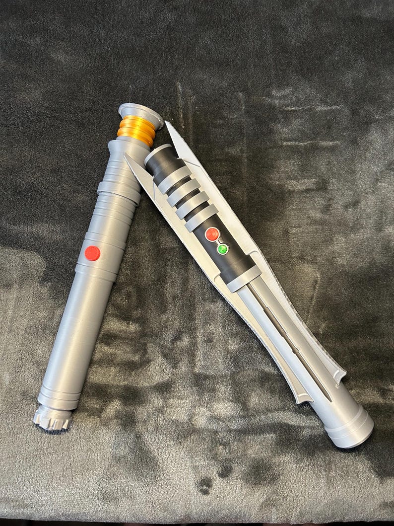 May include: Two toy lightsabers. One is silver with a red button and orange accents. The other is silver and black with a red and green button. The lightsabers are lying on a dark grey surface.