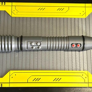 Op de afbeelding: Een zilveren lichtzwaard prop met een geribbelde handgreep en zwarte accenten. De prop heeft twee rode knoppen en twee kleinere knoppen. Het lichtzwaard ligt op een grijs oppervlak met gele en zwarte rand.