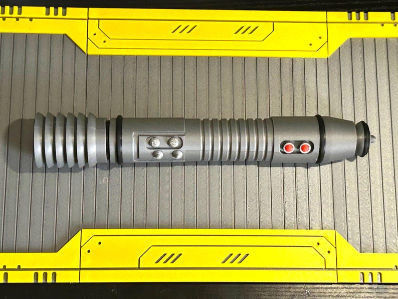 Op de afbeelding: Een zilveren lichtzwaard prop met een geribbelde handgreep en zwarte accenten. De prop heeft twee rode knoppen en twee kleinere knoppen. Het lichtzwaard ligt op een grijs oppervlak met gele en zwarte rand.