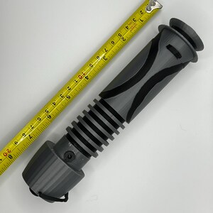 Peut inclure: Une poign&eacute;e de sabre laser grise avec une poign&eacute;e noire et une section nervur&eacute;e. La poign&eacute;e mesure environ 28 cm de long.