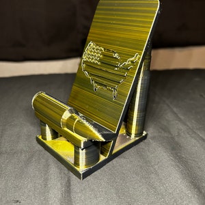 May include: A gold-colored phone stand with a 3D printed design. The stand features a silhouette of the United States with an American flag pattern and a bullet-shaped base.