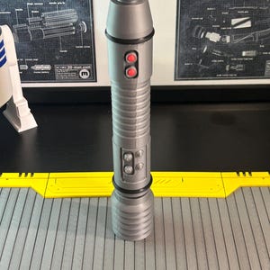 Op de afbeelding: Een zilveren lichtzwaard prop met rode knoppen en een gedetailleerd ontwerp. De prop is cilindrisch met een geribbelde handgreep en een zwart accent in de buurt van de bovenkant. De achtergrond bevat Star Wars schema's en een geel en grijs oppervlak.