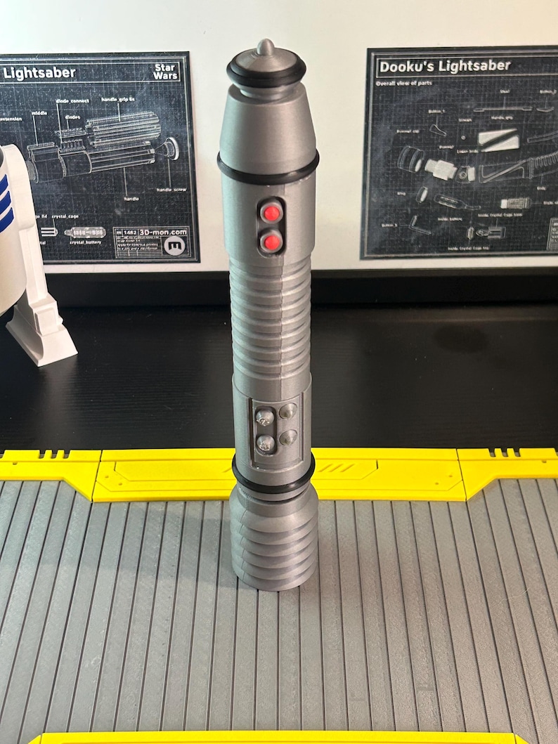 Op de afbeelding: Een zilveren lichtzwaard prop met rode knoppen en een gedetailleerd ontwerp. De prop is cilindrisch met een geribbelde handgreep en een zwart accent in de buurt van de bovenkant. De achtergrond bevat Star Wars schema's en een geel en grijs oppervlak.
