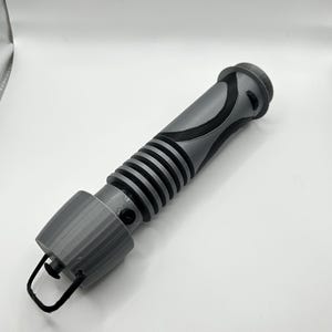 Peut inclure: Une r&eacute;plique de sabre laser imprim&eacute;e en 3D de couleur grise avec un motif noir et gris. Le sabre laser a une surface textur&eacute;e et une petite boucle &agrave; l'extr&eacute;mit&eacute;.