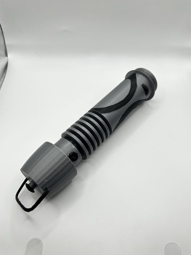 Peut inclure: Une r&eacute;plique de sabre laser imprim&eacute;e en 3D de couleur grise avec un motif noir et gris. Le sabre laser a une surface textur&eacute;e et une petite boucle &agrave; l'extr&eacute;mit&eacute;.