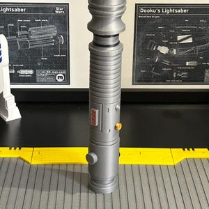 Op de afbeelding: Een grijze lichtzwaard prop met een cilindrisch lichaam, met een gedetailleerd ontwerp met ribbels, knoppen en een rood paneel. De prop wordt weergegeven op een grijs en geel oppervlak, met Star Wars schema's op de achtergrond.