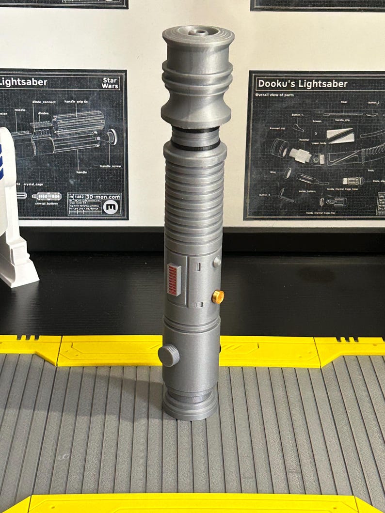 Op de afbeelding: Een grijze lichtzwaard prop met een cilindrisch lichaam, met een gedetailleerd ontwerp met ribbels, knoppen en een rood paneel. De prop wordt weergegeven op een grijs en geel oppervlak, met Star Wars schema's op de achtergrond.
