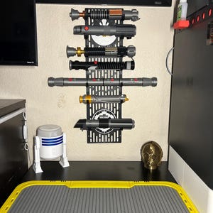 May include: A black wall-mounted display rack holding several lightsabers of various colors and designs. The rack is mounted on a beige wall, with a desk below holding a R2-D2 figurine, a gold-colored head, and a yellow and gray mat.