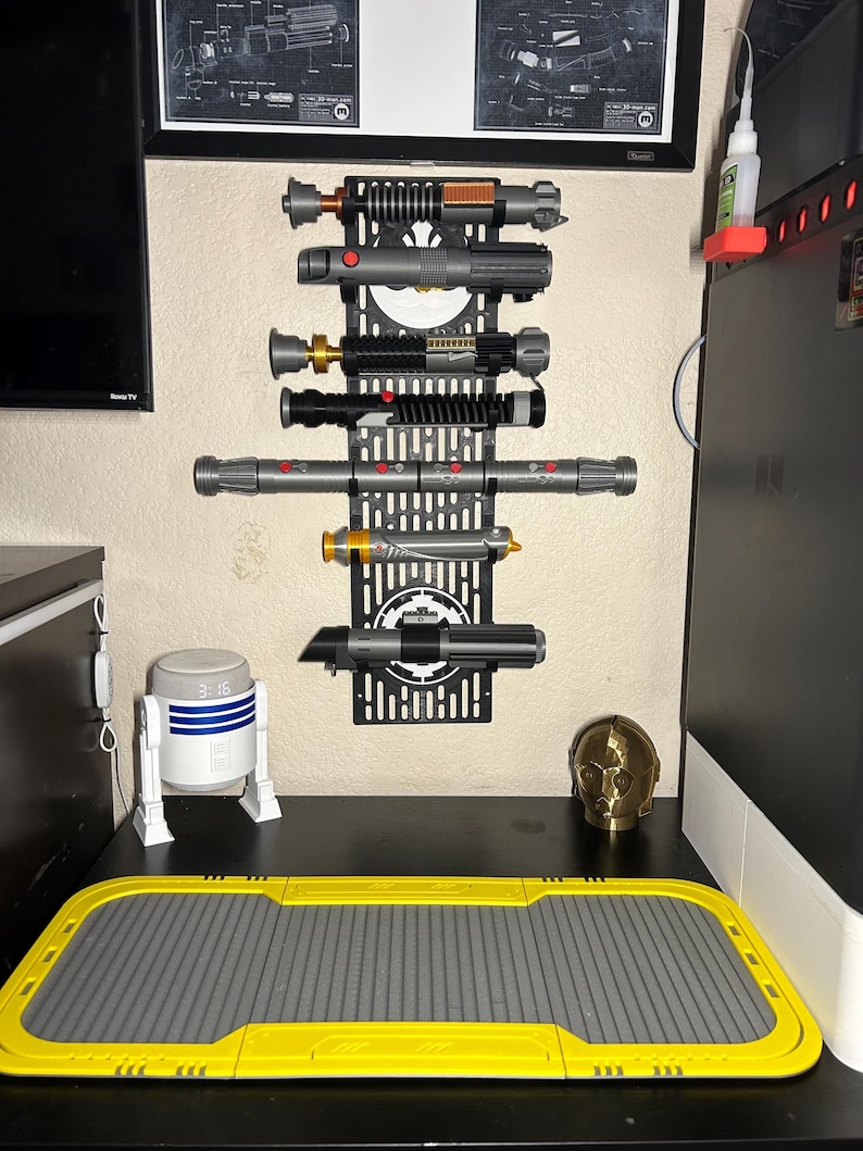 May include: A black wall-mounted display rack holding several lightsabers of various colors and designs. The rack is mounted on a beige wall, with a desk below holding a R2-D2 figurine, a gold-colored head, and a yellow and gray mat.