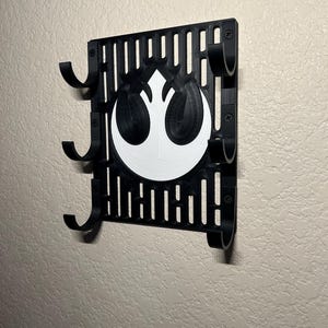 May include: A black wall-mounted organizer featuring a white Rebel Alliance symbol. The organizer has multiple curved hooks and a slotted design. It is mounted on a textured, light-colored wall.