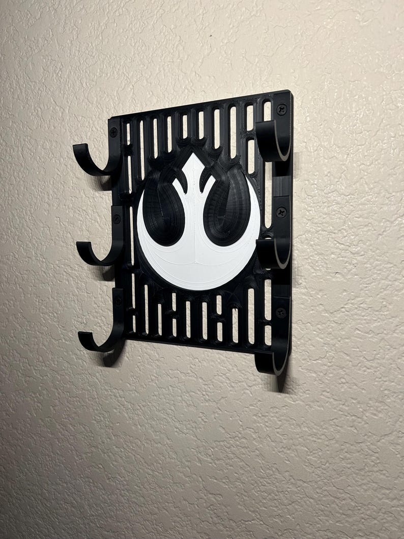 May include: A black wall-mounted organizer featuring a white Rebel Alliance symbol. The organizer has multiple curved hooks and a slotted design. It is mounted on a textured, light-colored wall.
