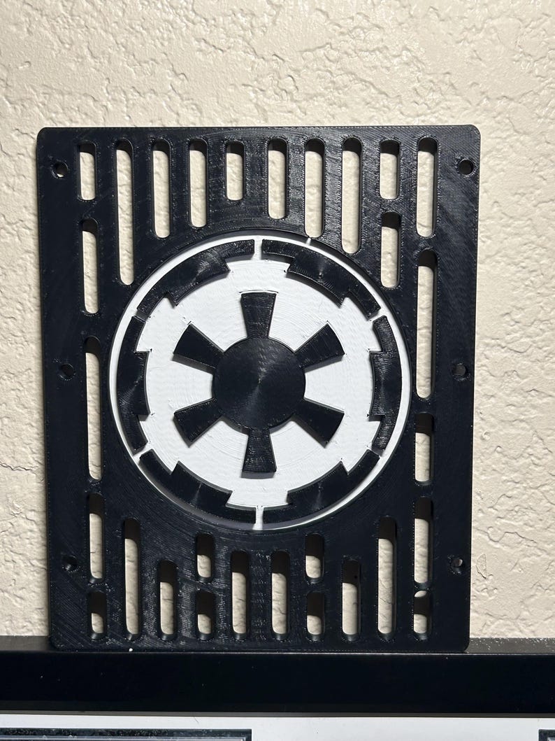 May include: Black and white 3D-printed wall art featuring the Galactic Empire symbol. The symbol is centered within a rectangular frame with vertical cutouts. The design is a fan-made item.