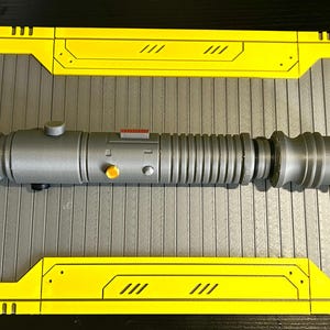 Op de afbeelding: Een zilveren lichtzwaard prop met een cilindrisch ontwerp, met geribbelde secties en een afgeronde pommel. Het heeft oranje en rode accenten en een kleine knop. Het lichtzwaard ligt op een grijze ondergrond met gele panelen.