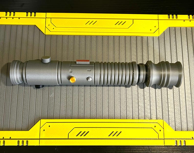 Op de afbeelding: Een zilveren lichtzwaard prop met een cilindrisch ontwerp, met geribbelde secties en een afgeronde pommel. Het heeft oranje en rode accenten en een kleine knop. Het lichtzwaard ligt op een grijze ondergrond met gele panelen.