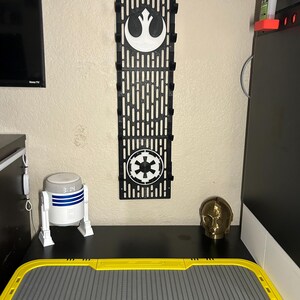 May include: A black wall organizer featuring the Rebel Alliance and Galactic Empire symbols. A white and blue R2-D2 clock, a gold C-3PO head, and a yellow and gray tray are on a black surface.