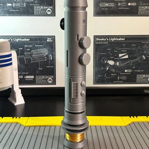 Op de afbeelding: Een grijze lichtzwaard prop met een cilindrisch lichaam, ronde knoppen en een gedetailleerd onderste gedeelte. Het handvat heeft een goudkleurige basis en een zwarte knop. Achtergrond bevat Star Wars schema's.