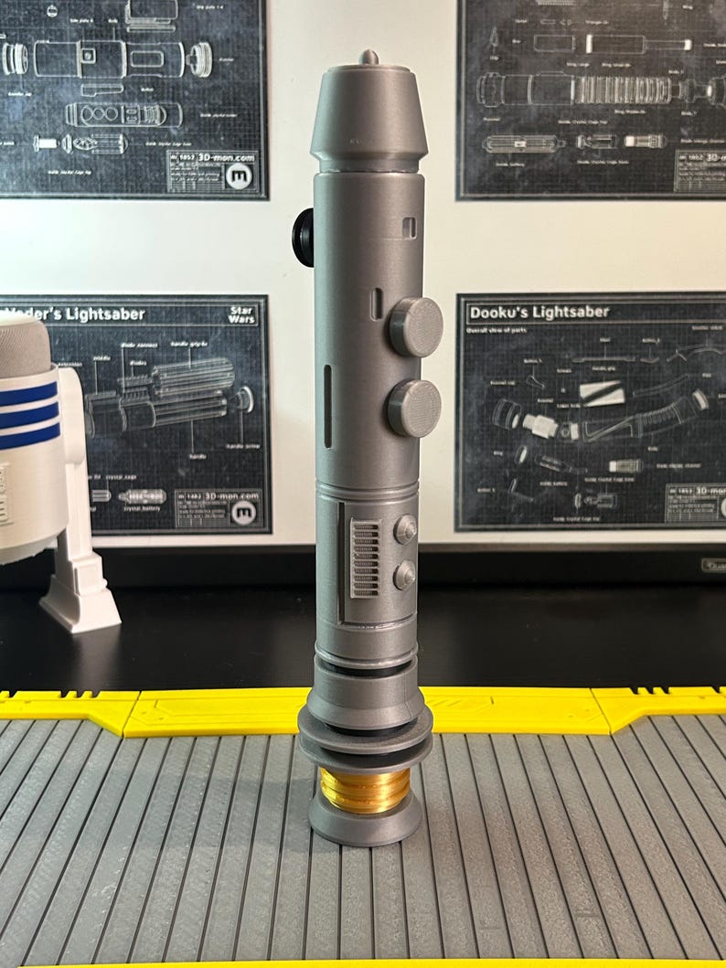 Op de afbeelding: Een grijze lichtzwaard prop met een cilindrisch lichaam, ronde knoppen en een gedetailleerd onderste gedeelte. Het handvat heeft een goudkleurige basis en een zwarte knop. Achtergrond bevat Star Wars schema's.