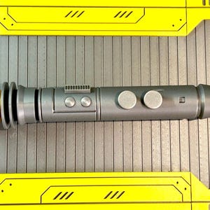 Op de afbeelding: Een zilveren en gouden lichtzwaard prop met een cilindrisch lichaam en gedetailleerd ontwerp. De prop heeft twee zilveren knoppen en een goudkleurige emitter. Het lichtzwaard wordt weergegeven op een grijze ondergrond met gele accenten.