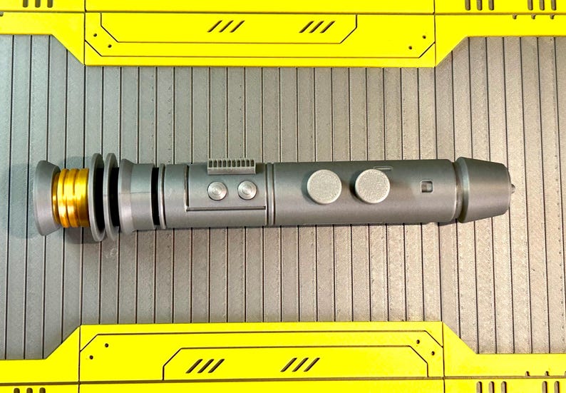 Op de afbeelding: Een zilveren en gouden lichtzwaard prop met een cilindrisch lichaam en gedetailleerd ontwerp. De prop heeft twee zilveren knoppen en een goudkleurige emitter. Het lichtzwaard wordt weergegeven op een grijze ondergrond met gele accenten.