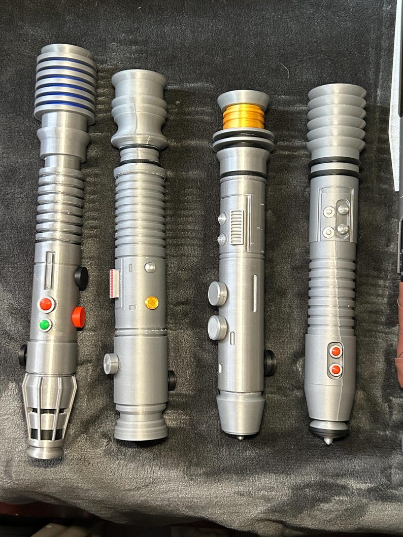 Op de afbeelding: Vier zilverkleurige lichtzwaardreplica's met gedetailleerde ontwerpen. De lichtzwaarden hebben verschillende knop- en gripconfiguraties, met accenten van blauw, oranje en rood. De lichtzwaarden zijn gerangschikt op een donkergrijze ondergrond.