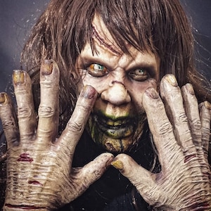 May include: A close-up of a person with a scary, grotesque makeup look. They have green and black makeup around their mouth, and their eyes are wide open with a creepy expression. They are wearing a dark shirt and have their hands up in front of their face.